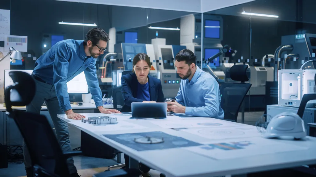 Three people in a modern office sitting around a table with a laptop, reviewing documents and technical drawings, with machinery and monitors visible in the background.