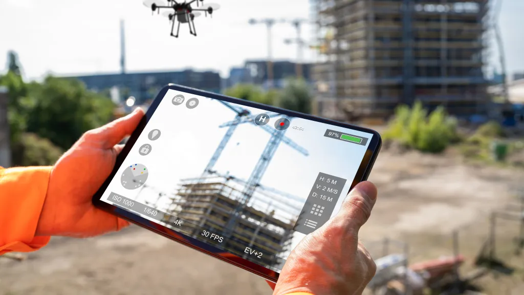 Worker using a tablet to pilot a drone over a construction site, with cranes and a multi-storey building in the background.