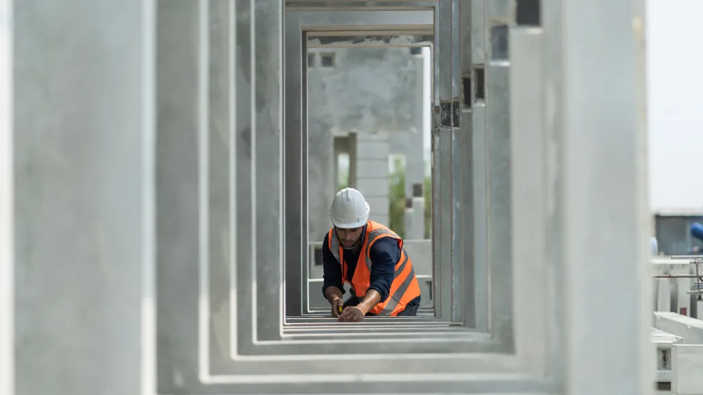 "Person wearing a hard hat and orange safety vest working with a measuring tool inside a series of concrete rectangular frames at a construction site.