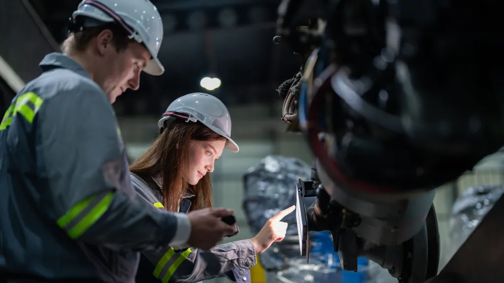 Two people wearing safety helmets and reflective clothing inspecting industrial machinery, with one person pointing at a control panel.