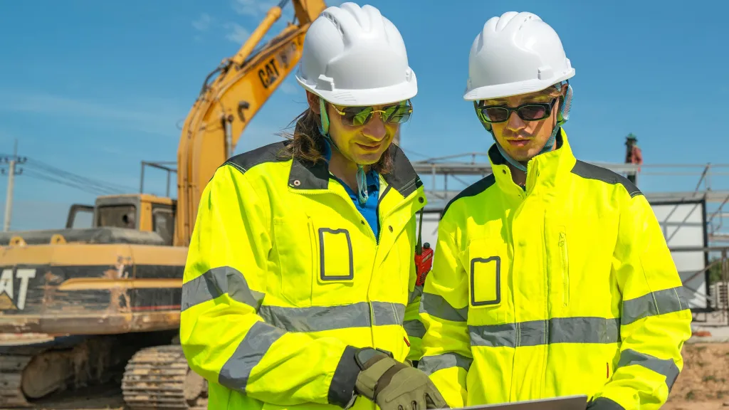 Two construction workers in hard hats and high-vis jackets reviewing plans at a building site.