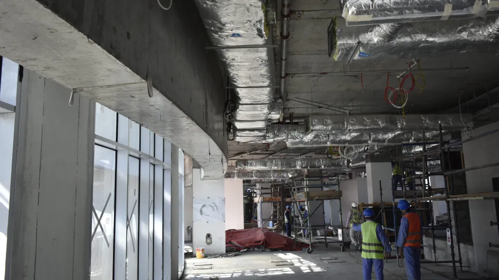 Construction site interior showing exposed ceiling with pipes and ductwork, and unfinished concrete walls.