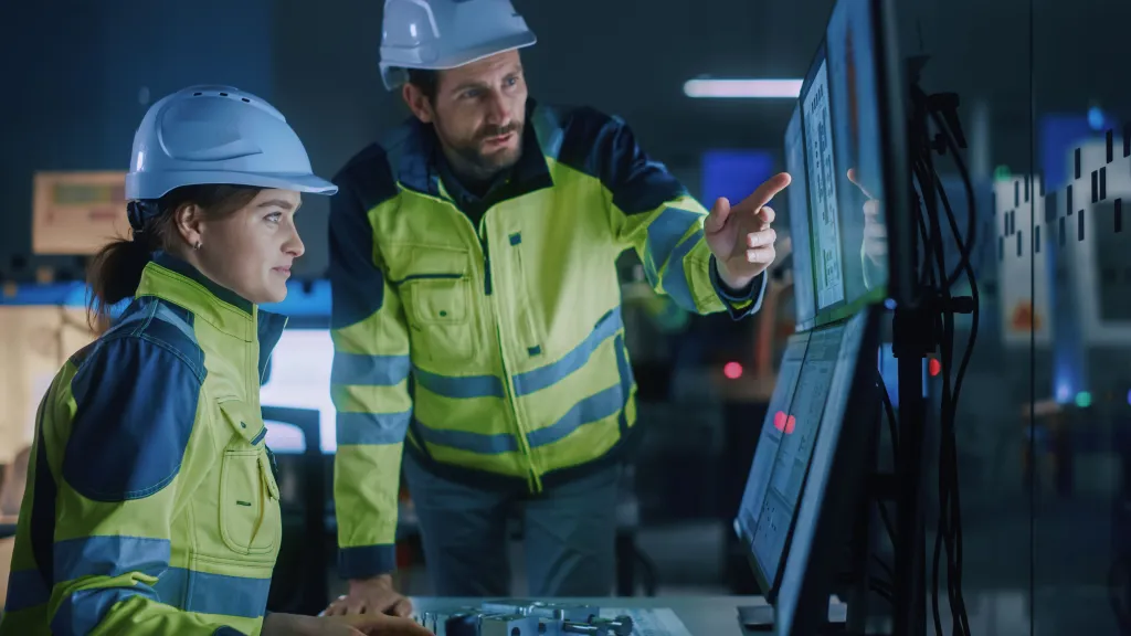 Two workers in hard hats and safety vests examining a digital screen in an industrial setting.