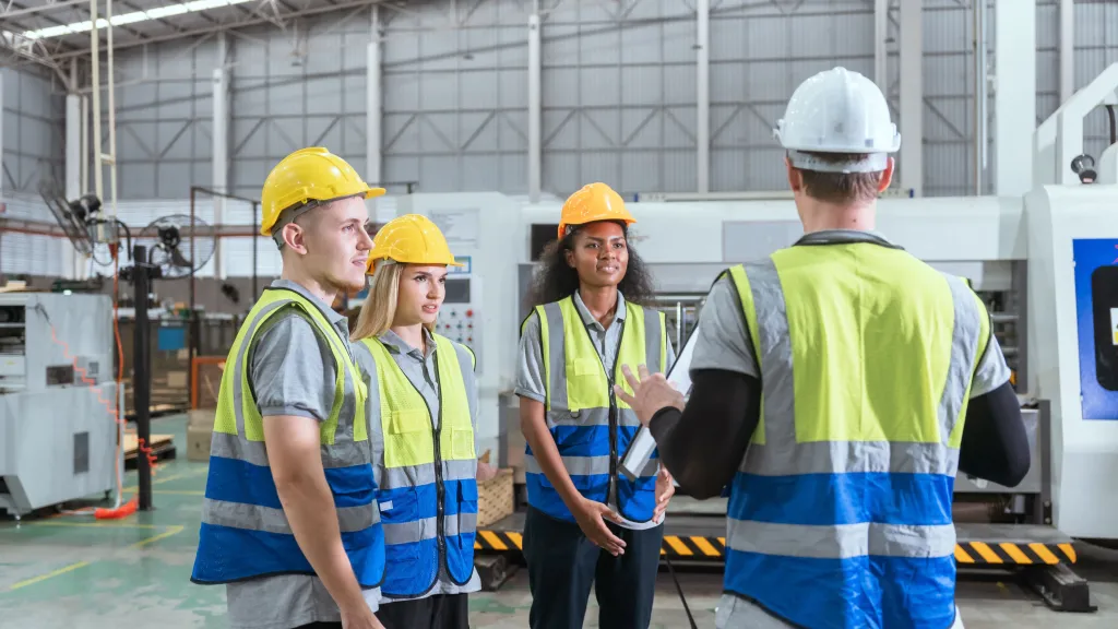 Three workers in hard hats, safety vests, and protective gear discussing at an industrial facility.
