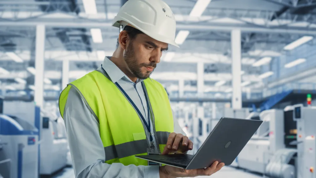 Worker in hard hat and safety vest using laptop in industrial facility.