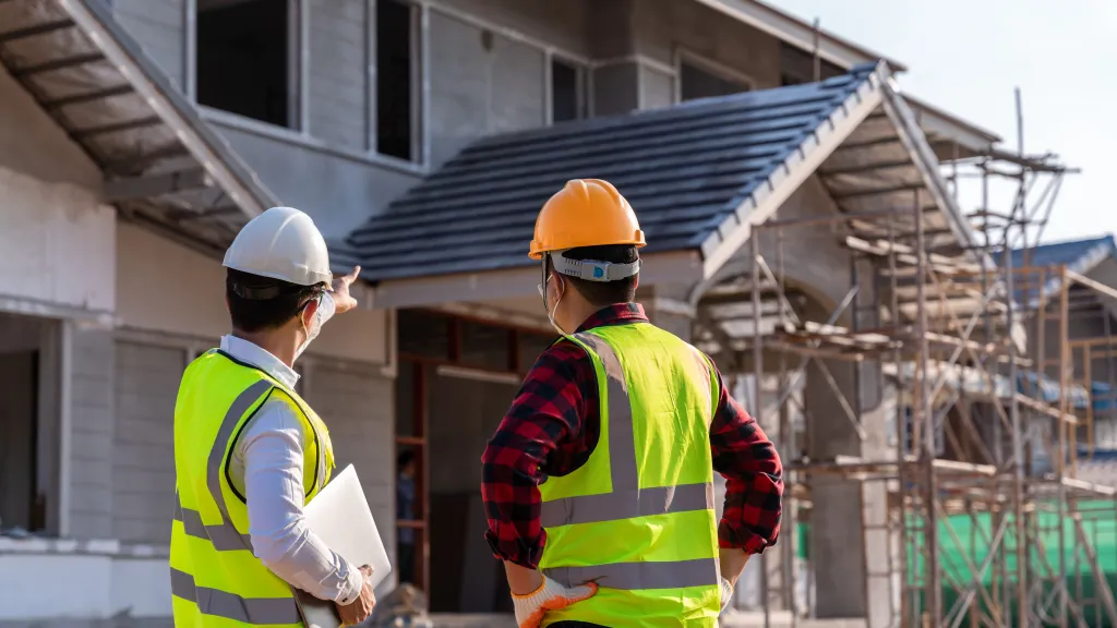 Two construction workers in hard hats and safety vests observing a residential building under construction.