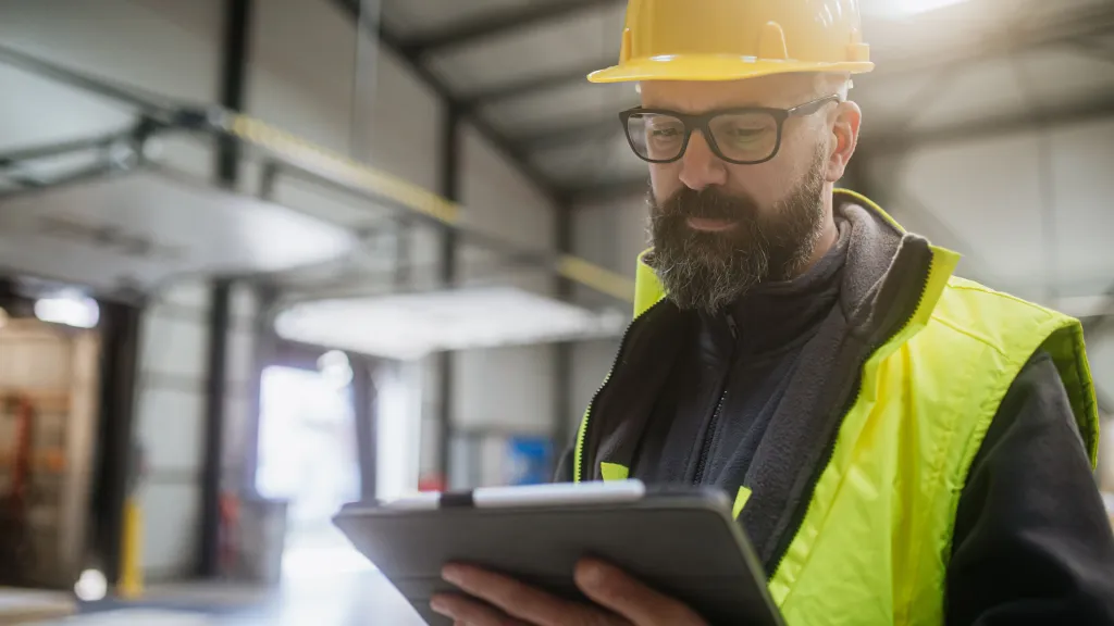 Construction worker in yellow hardhat and safety vest reviewing information on a tablet.