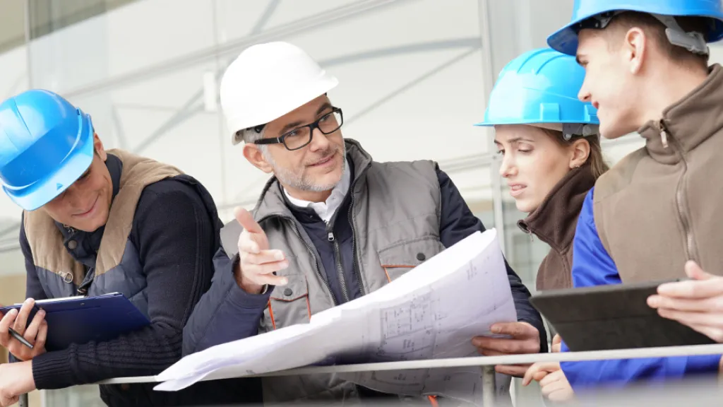 Construction professionals in blue hard hats examining blueprints at a worksite.
