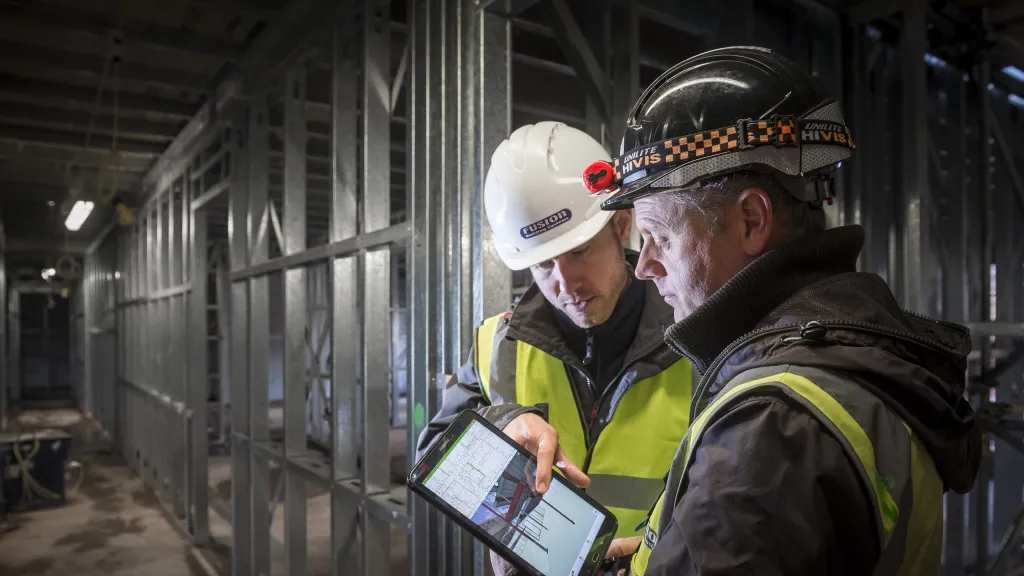 Two construction workers in safety gear examining a document inside an unfinished building.