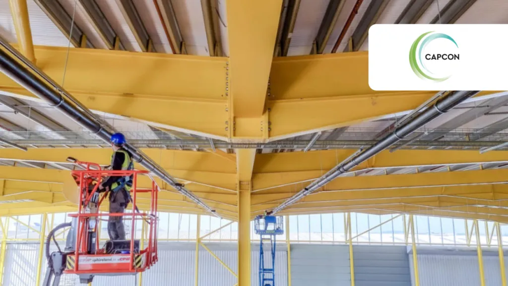 Worker on a yellow aerial lift platform inspecting a industrial building's ceiling structure.