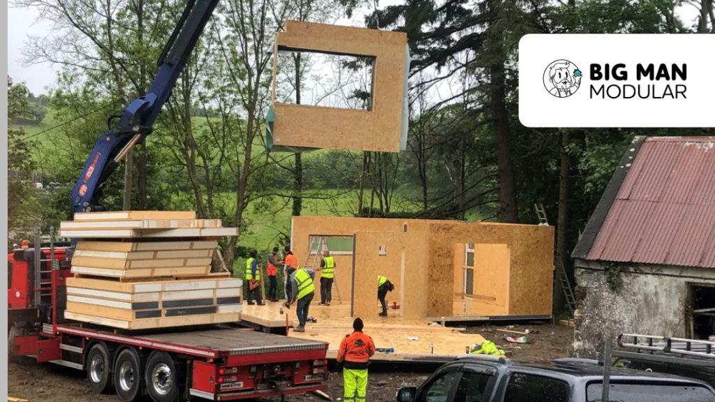 A crane lifts a modular wooden wall panel onto a construction site, with workers guiding it into place.