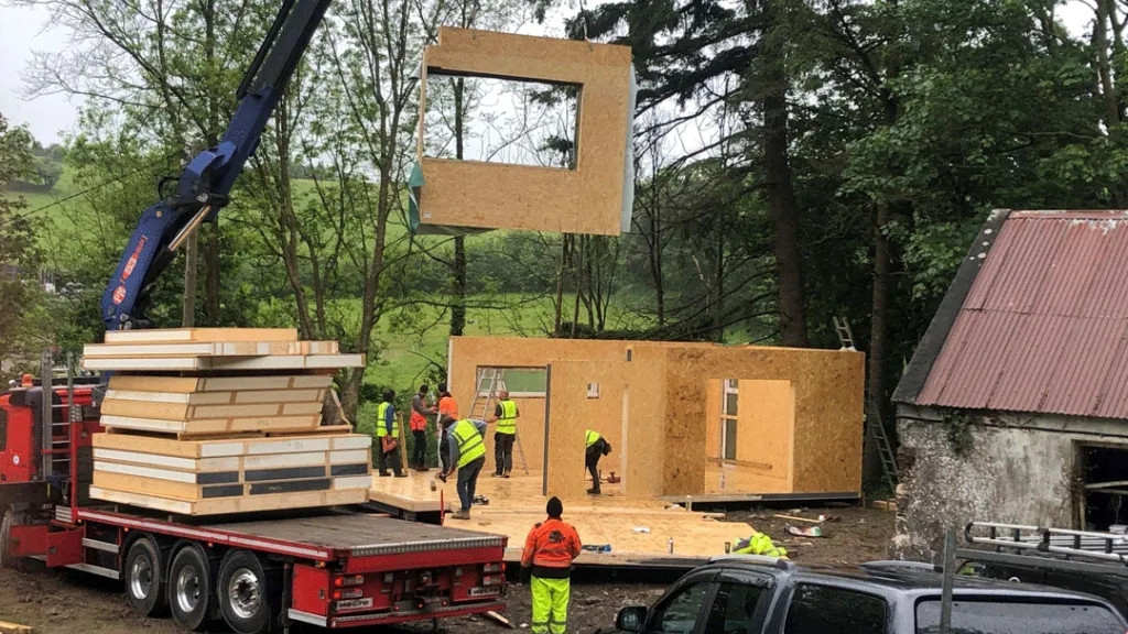 A crane lifts a large wooden frame module onto a modular home construction site surrounded by trees.