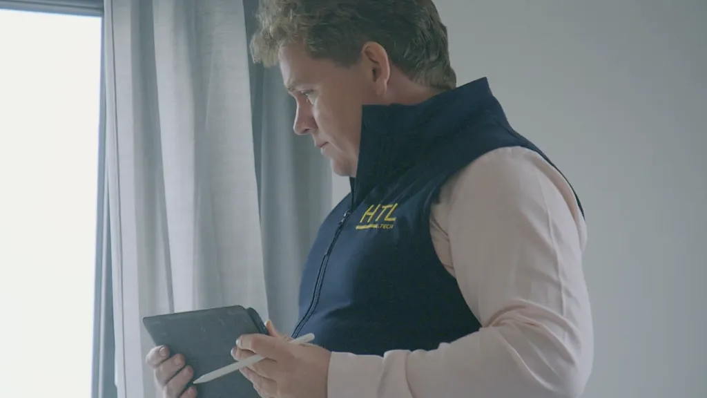 Man in a navy vest holding a tablet near a window indoors.