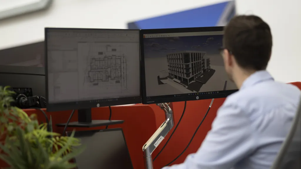 Man in blue shirt working at dual monitor workstation displaying CAD designs.