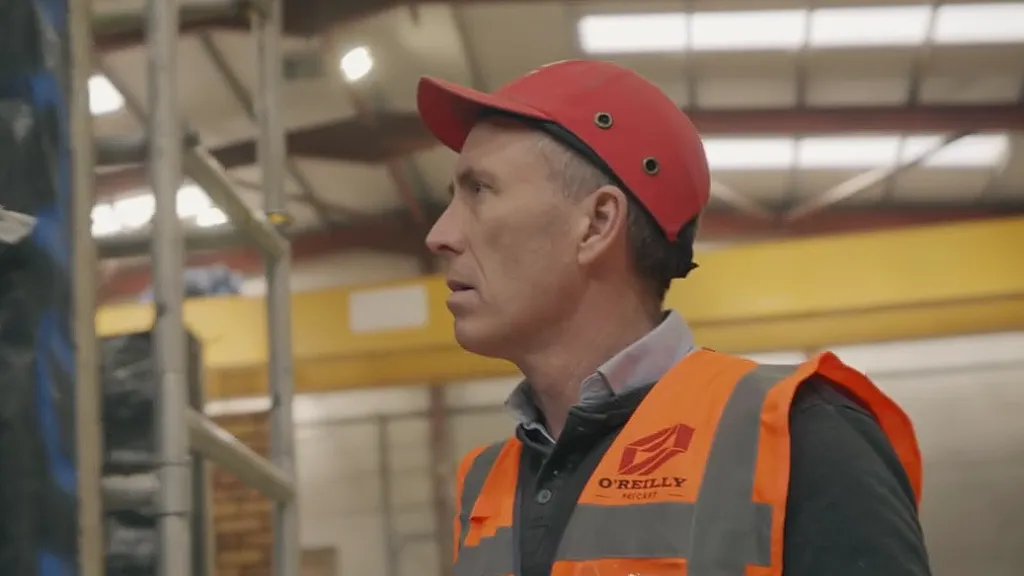 Man in orange safety vest and red hard hat looking sideways in an industrial warehouse.
