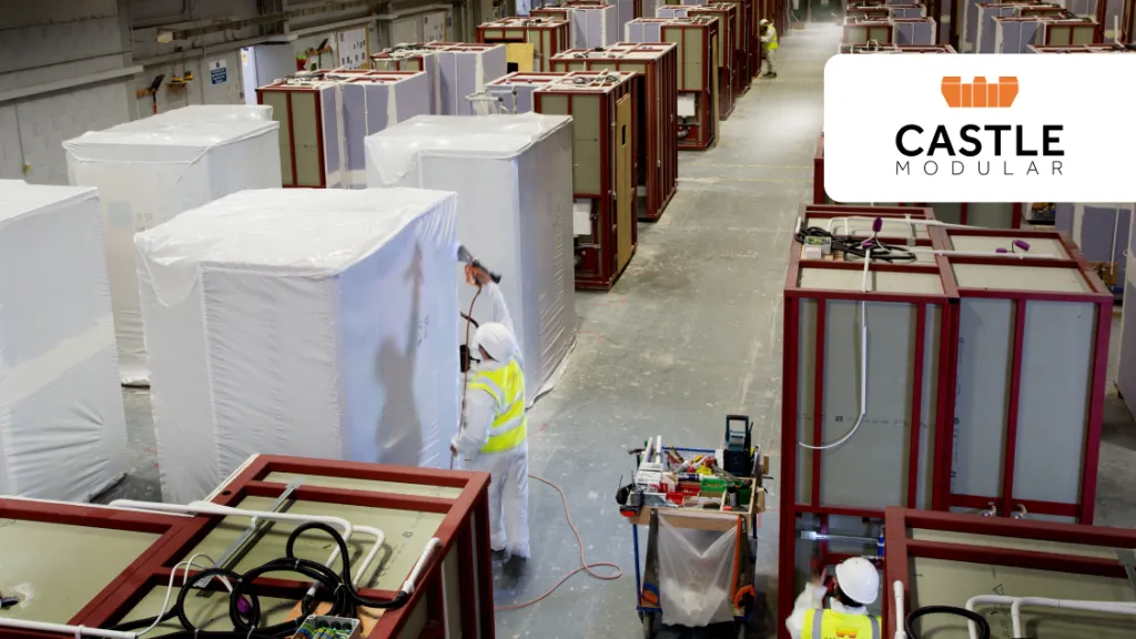 Factory workers preparing prefabricated construction modules inside a large manufacturing facility. Numerous partially assembled modules are arranged throughout the factory floor.