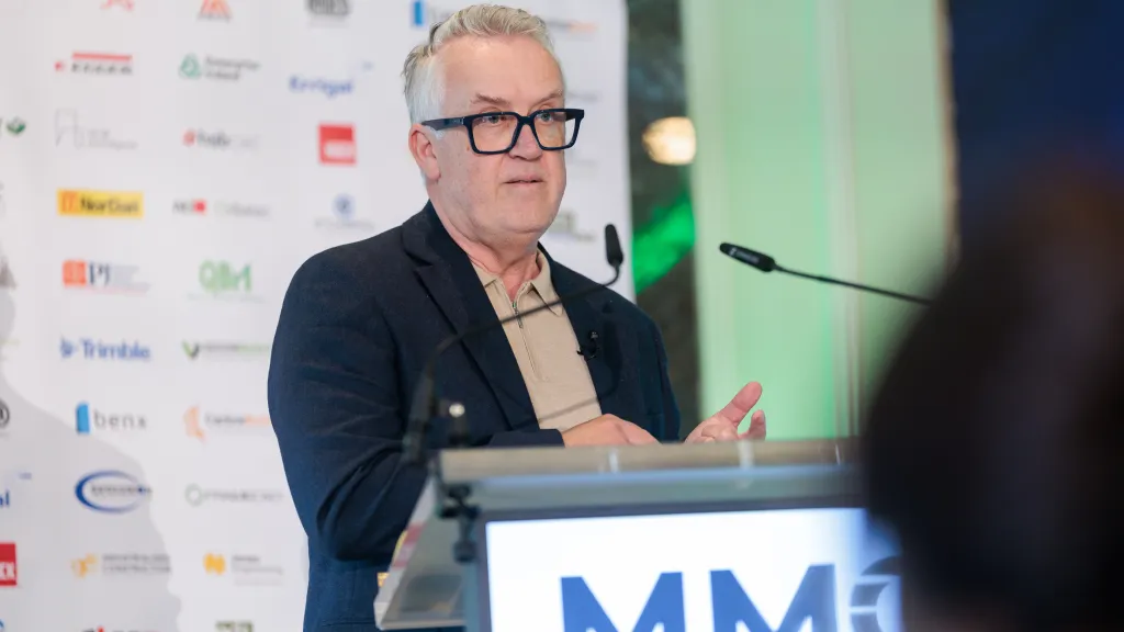 Older man with glasses speaking at a podium labeled "MM" at a formal event.