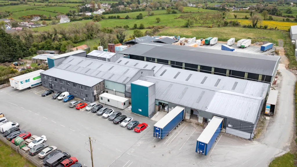 Aerial view of a large industrial warehouse complex with trucks and vehicles parked outside, surrounded by green countryside.