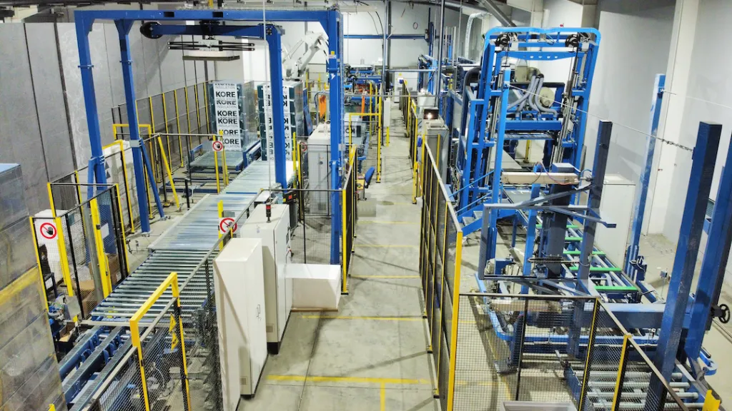 Industrial manufacturing facility with blue metal frames, conveyor systems, and white panels on the floor.