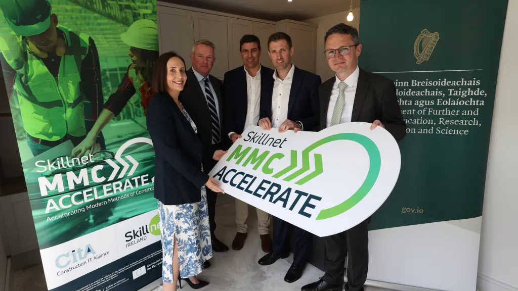 Five professionals posing together, holding a green "MMC Accelerate" sign at a formal event.