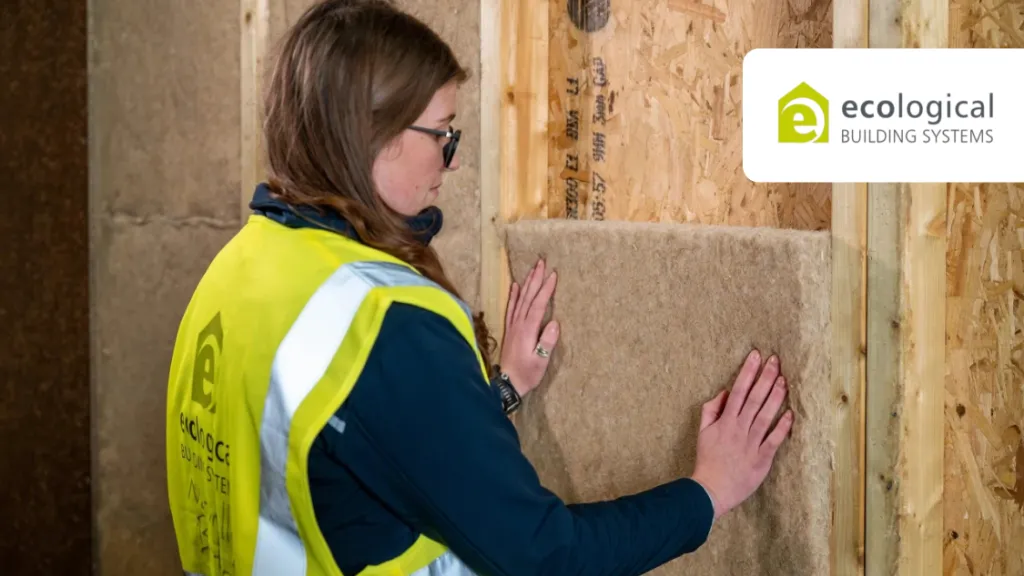 Woman in yellow safety vest installing mineral wool insulation panels in a wooden wall frame.