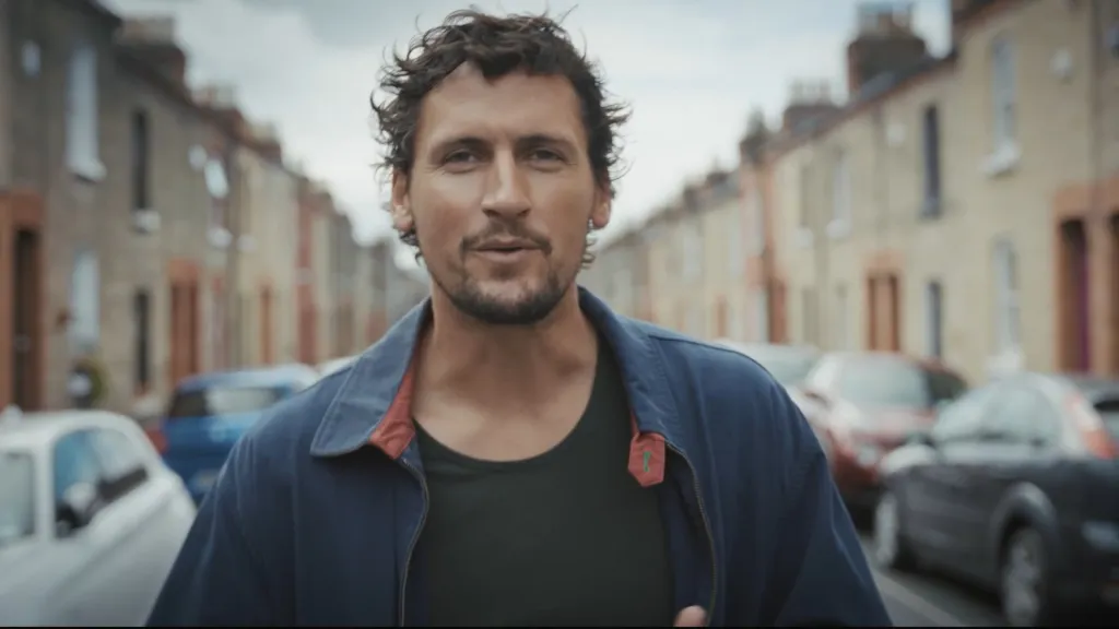 Man in blue jacket standing on a quiet residential street, looking directly at camera.
