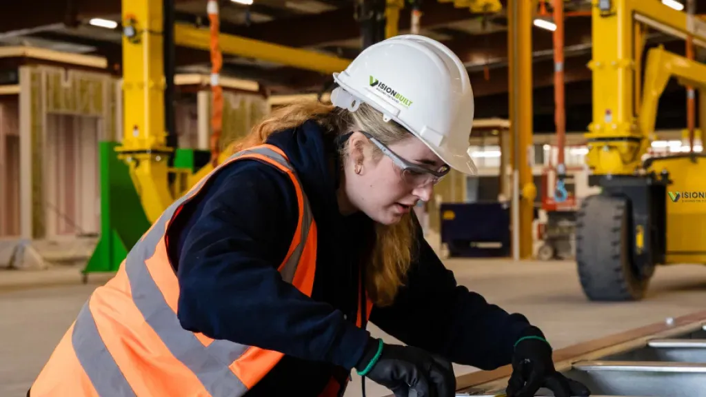 Worker in white hard hat and orange vest focused on task in industrial warehouse setting.