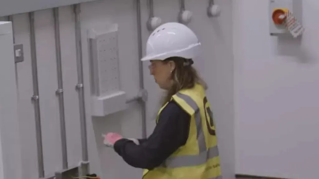 Woman in hard hat and yellow safety vest inspecting industrial equipment indoors.