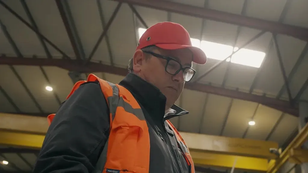 Worker in orange safety vest and red hard hat looking down in an industrial facility.