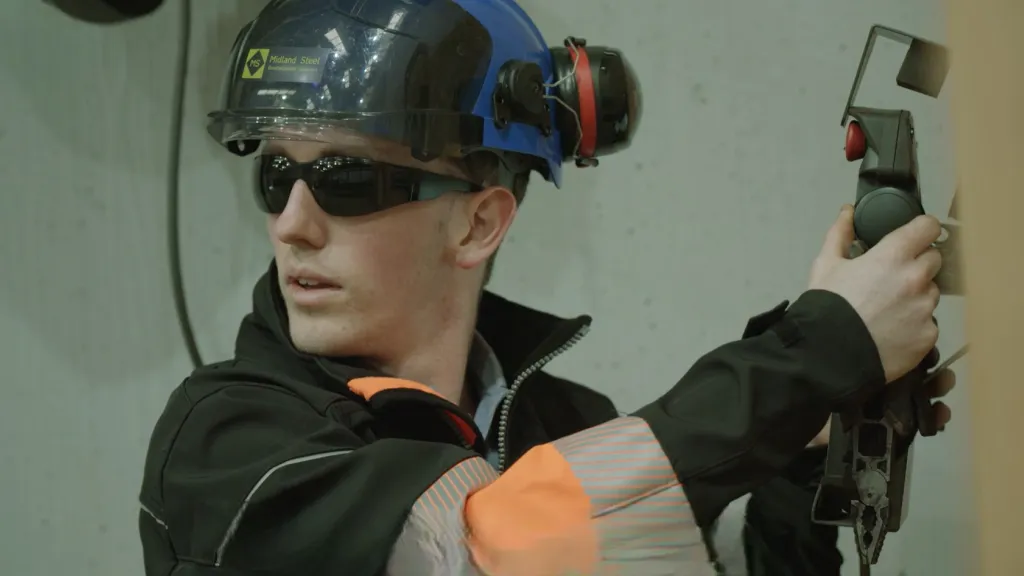 Person in protective gear with welding helmet and blue ear muffs, holding equipment indoors.
