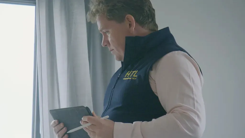 Man in a navy vest using a tablet near a bright window indoors.