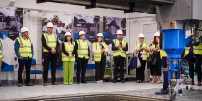 A group of people wearing high-visibility vests and hard hats standing in a row inside an industrial or construction facility, with machinery visible in the foreground.