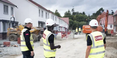 Three construction professionals in safety gear and hard hats are standing on a residential building site, discussing progress. Houses under construction are visible in the background.