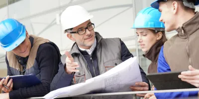 Construction professionals in blue hard hats examining blueprints at a worksite.