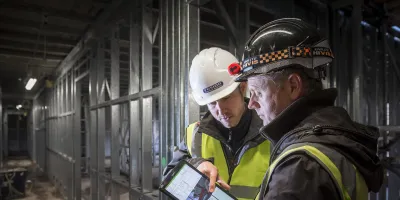 Two construction workers in safety gear examining a document inside an unfinished building.