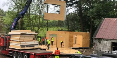 A crane lifts a large wooden frame module onto a modular home construction site surrounded by trees.