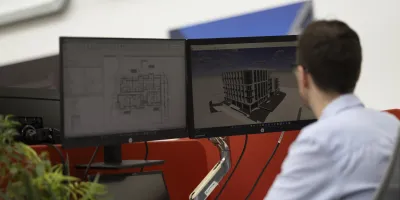 Man in blue shirt working at dual monitor workstation displaying CAD designs.
