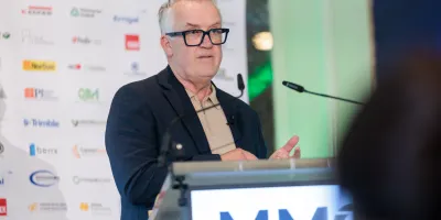 Older man with glasses speaking at a podium labeled "MM" at a formal event.