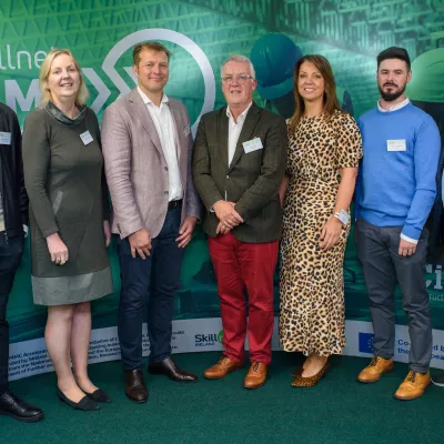 A group of people in smart clothing stand in front of a green screen with the Skillnet MMC Accelerate logo.