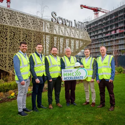 A group of males in high vis vests stand in front of a modern structure, holding a board has the Skillnet MMC Accelerate logo.