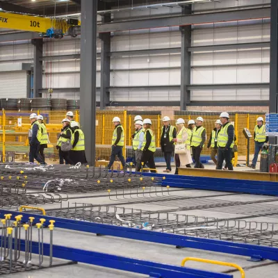 A group of people in high-vis jackets and white hard hats inspect a steel welding manufacturing facility.