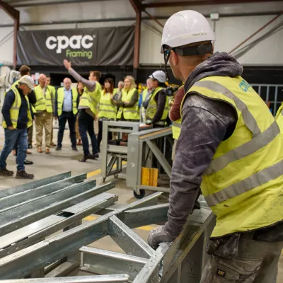 A man in a white hard hat and high vis vest watches a group of people listening to a man talking in the background
