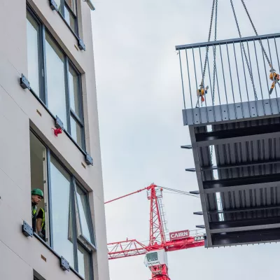 A male in a hard hat and high vis jacket watches a crane manoeuvre a metal balcony towards an apartment building.
