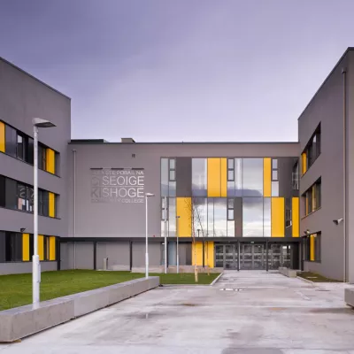 A modern grey school building with yellow and grey panels, surrounding a grass courtyard.