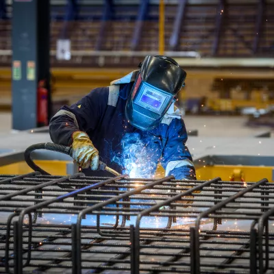 A person in protective clothing and a welding helmet uses a blowtorch to weld steel bars in a manufacturing facility.
