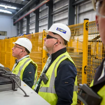 Three people in high-vis vests and hard hats observing machinery inside a construction factory.