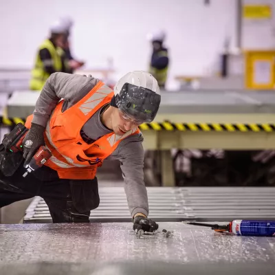 A male in a high vis jacket and hard hat holding a power tool crouches over a pallet.