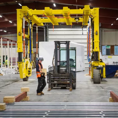 A person wearing a high vis vest and hard hat carries a wooden block through a manufacturing facility passed a forklift.
