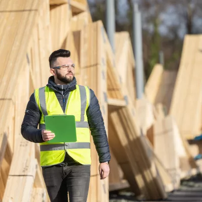 A male with protective goggles and a high-vis jacket carrying a clipboard passed piles of lumber. 