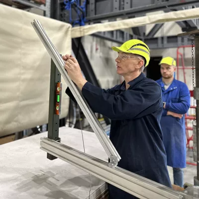 Two people in blue work overalls and hard hats measure a metal beam.